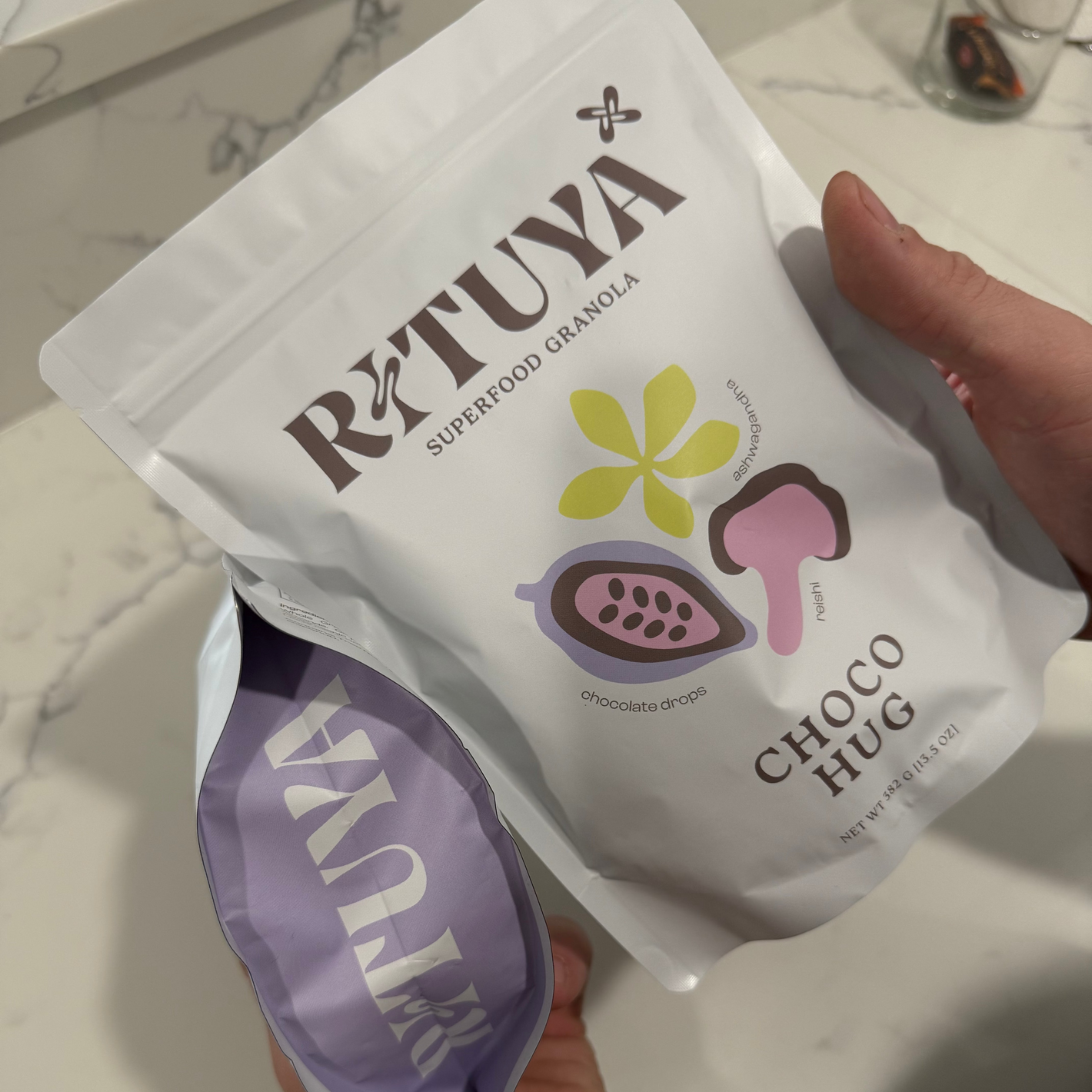 Two Rituva superfood granola packages, one white and one purple, held by a hand on a marble surface.